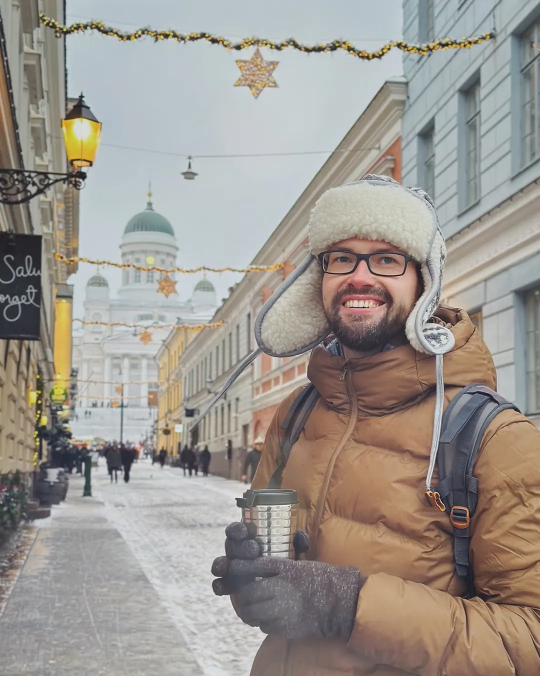 Andrey the Nomad on a Helsinki street in December, in a fur hat with a takeaway coffee, Helsinki Cathedral behind him