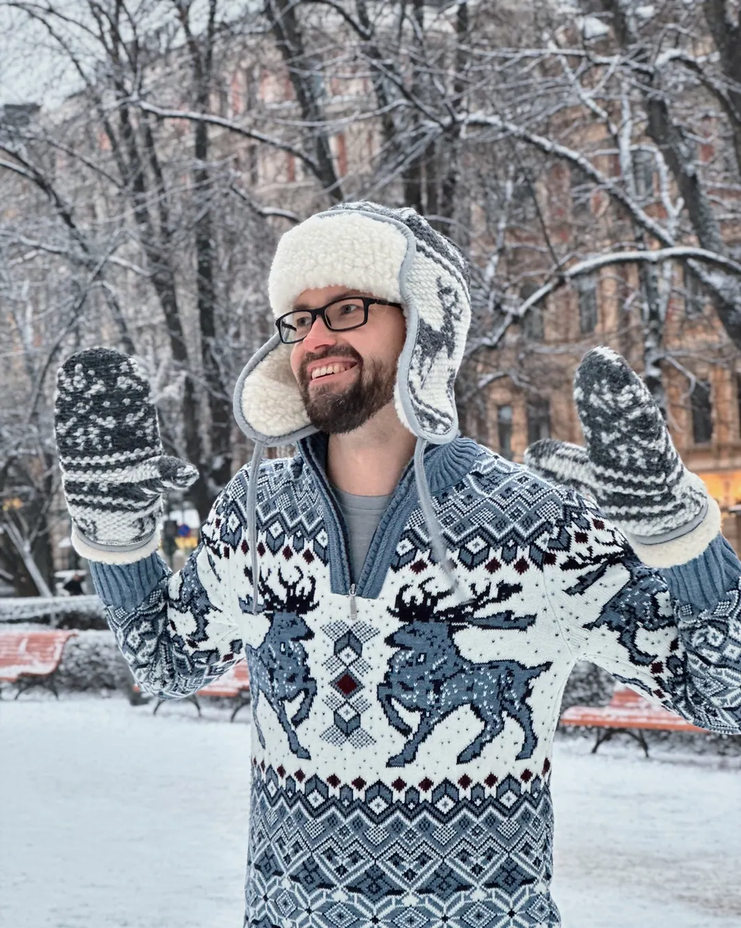 Andrey the Nomad in a knitted reindeer sweater and fur hat, mittens up, in a snowy Helsinki park