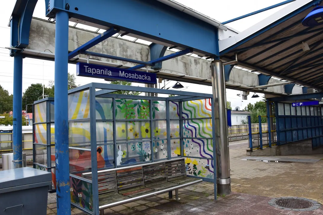 A blue station name board reading Tapanila Mosabacka above a painted glass shelter on a suburban platform