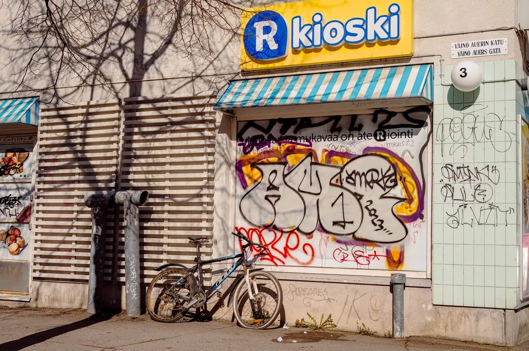 A shuttered R-kioski covered in graffiti, a bicycle leaning against it, and a street plate reading Väinö Auerin katu and Väinö Auers gata