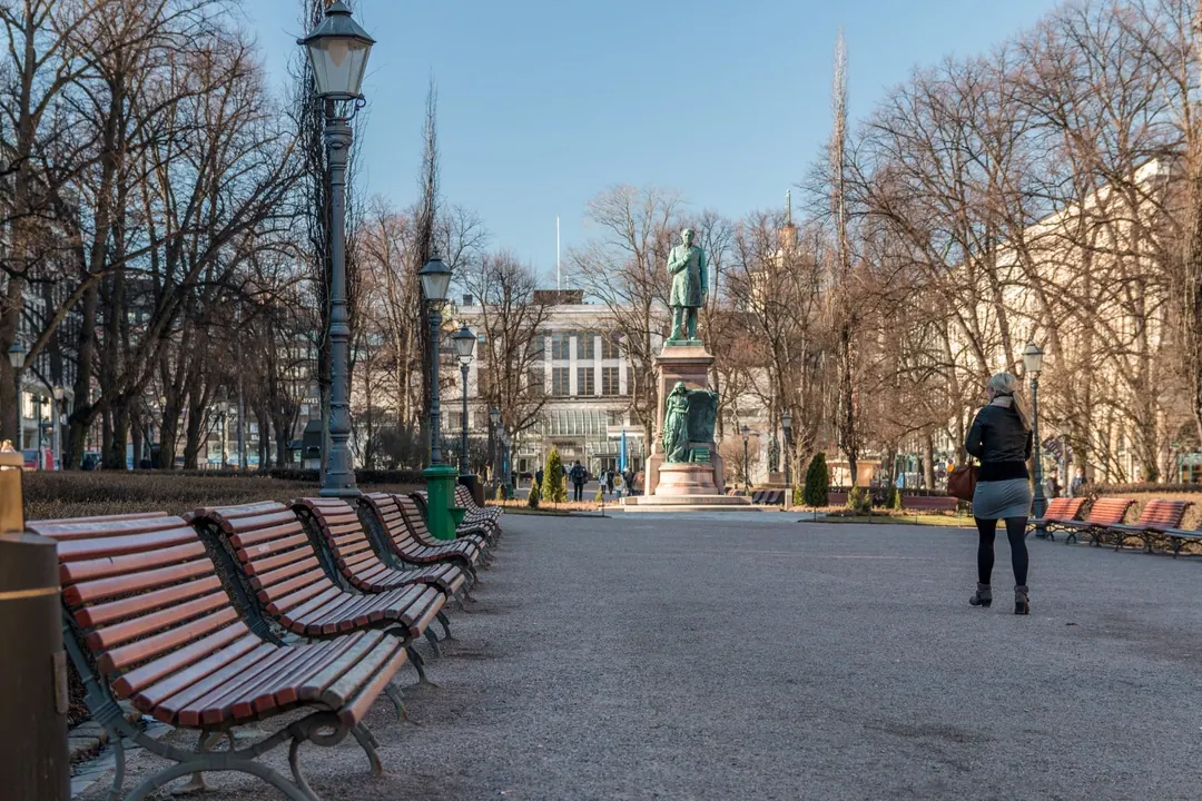 Bare trees and empty benches along Esplanadi park in early spring, one woman walking towards the Runeberg statue