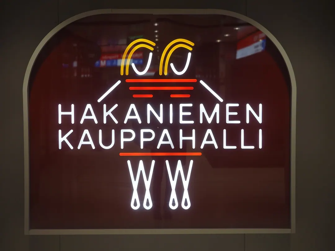The illuminated neon sign of Hakaniemen Kauppahalli, white lettering under an orange and yellow motif