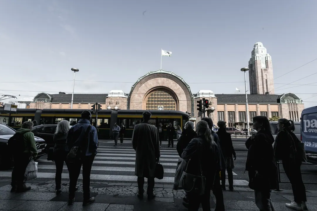 A crowd waiting at a crossing in front of Helsinki Central Station, a tram passing, the clock tower behind