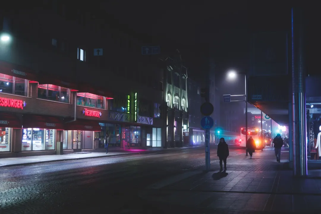 A misty night street in Oulu, neon shopfronts, one person waiting alone at a stop
