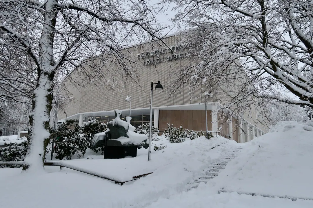Töölö library in Helsinki under deep snow, bare trees and a sculpture in the foreground, the name lettered across the facade