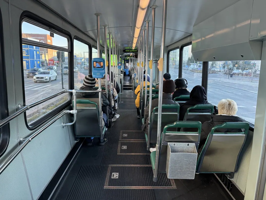 Inside a Helsinki tram in winter, passengers in hats seated facing forward, low sun through the windows