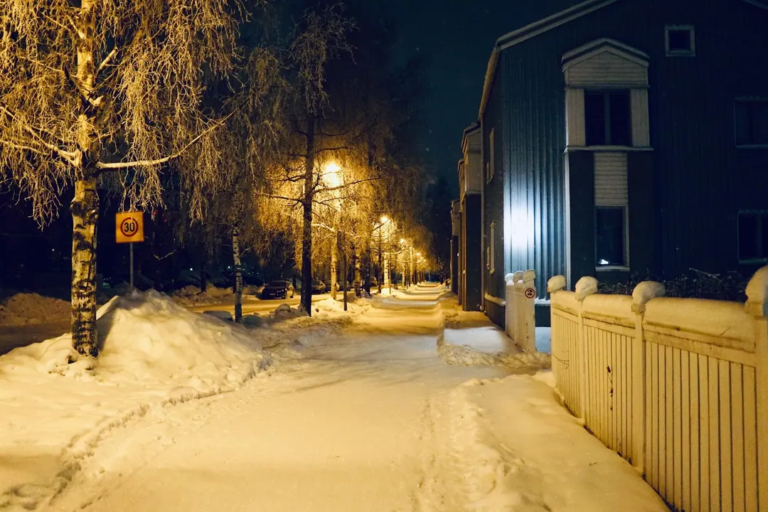 A snowy residential street in Oulu at night, street lamps lit, a dark timber house with one bright window