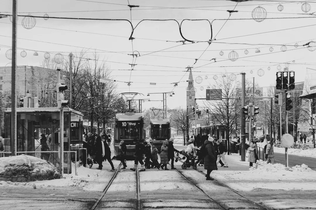A snowy Helsinki tram stop in black and white, people crossing the tracks in front of a tram, festive lights strung overhead