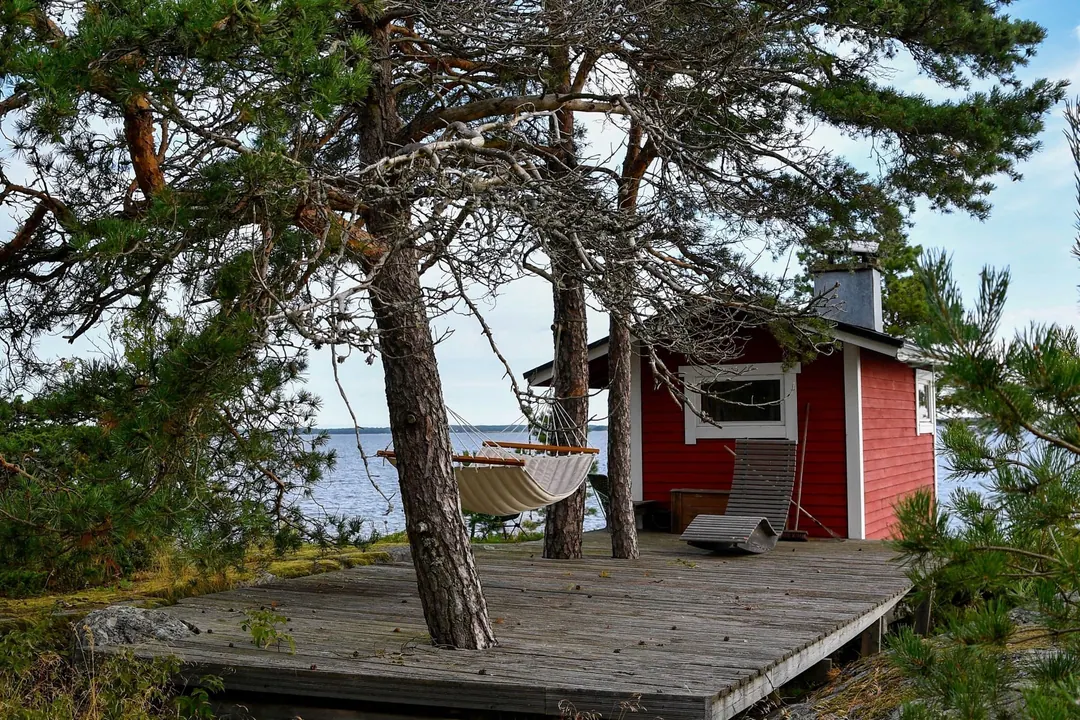 A red wooden cottage with a sauna chimney, a hammock and a plank deck at the water's edge
