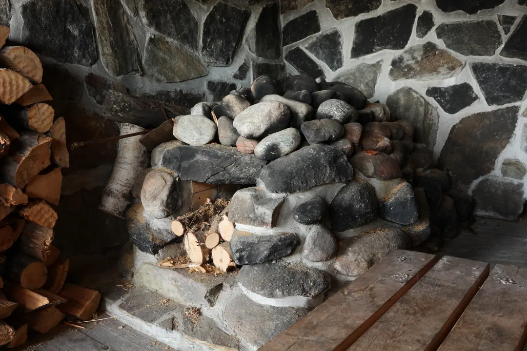 A smoke sauna stove built of loose stones on a stone hearth, split firewood stacked beside it