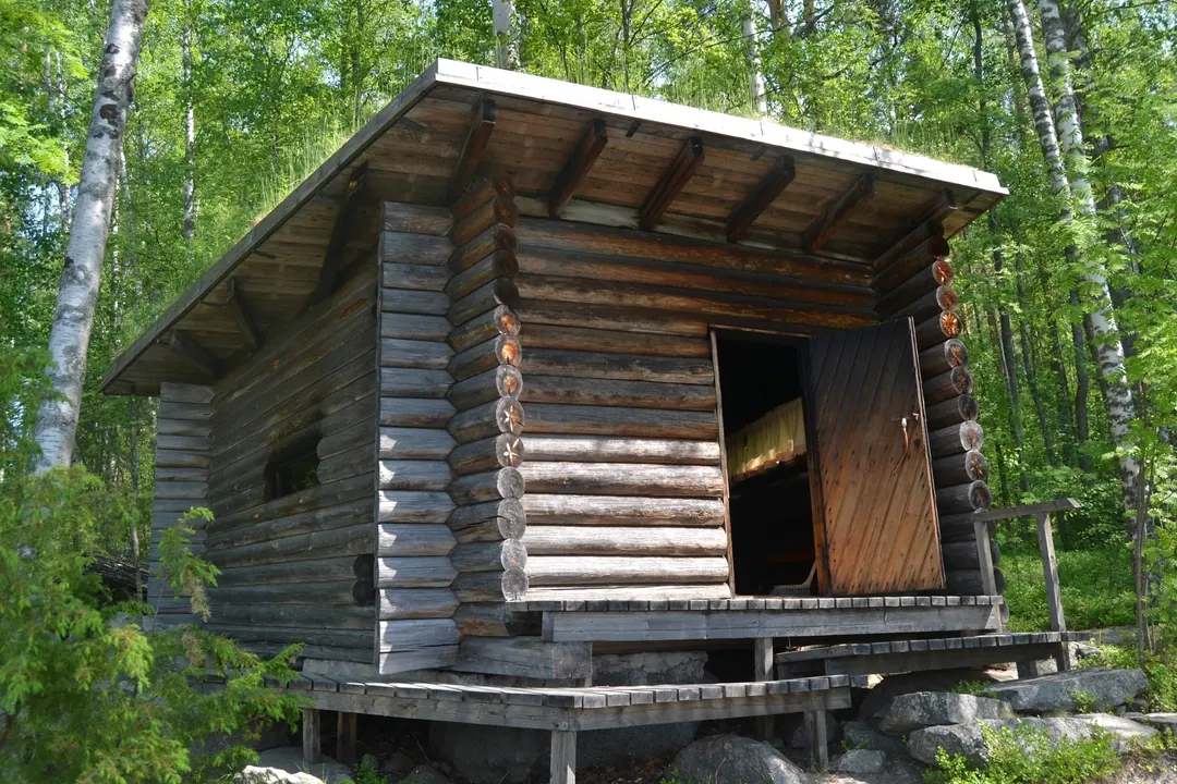 A small log smoke sauna standing on stones among birches beside a lake in summer