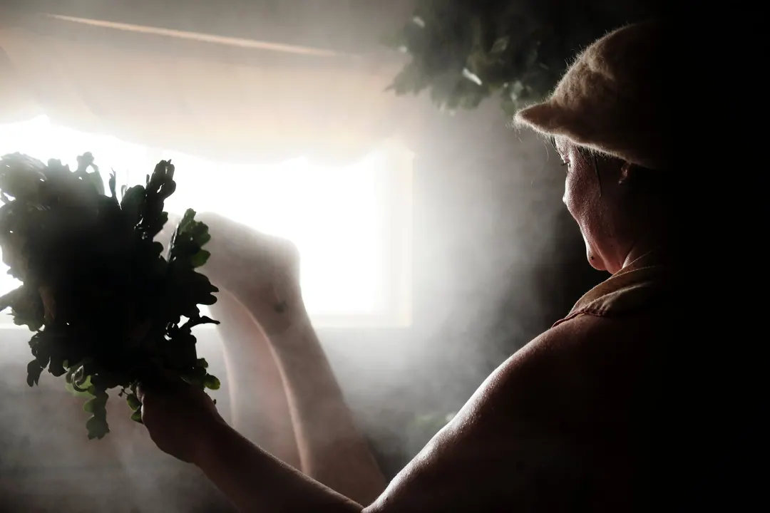 A woman in a felt sauna hat holding an oak whisk in a room full of steam