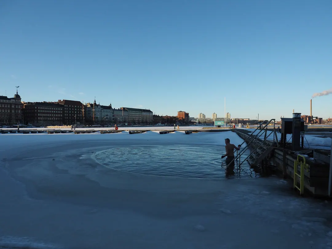 A man on a ladder in a hole cut in the sea ice at Katajanokka in Helsinki, apartment blocks across the water