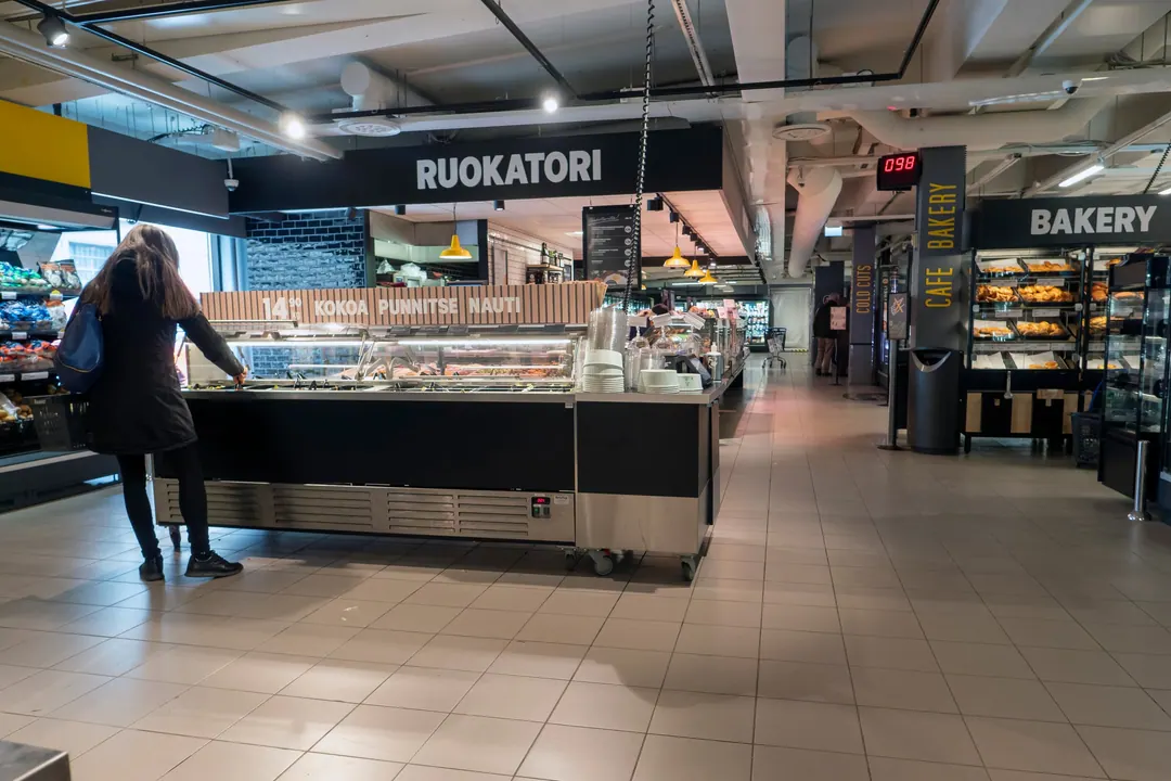 Inside a Helsinki S-market, a red queue-number display reading 098 above a counter, Finnish and English signage side by side