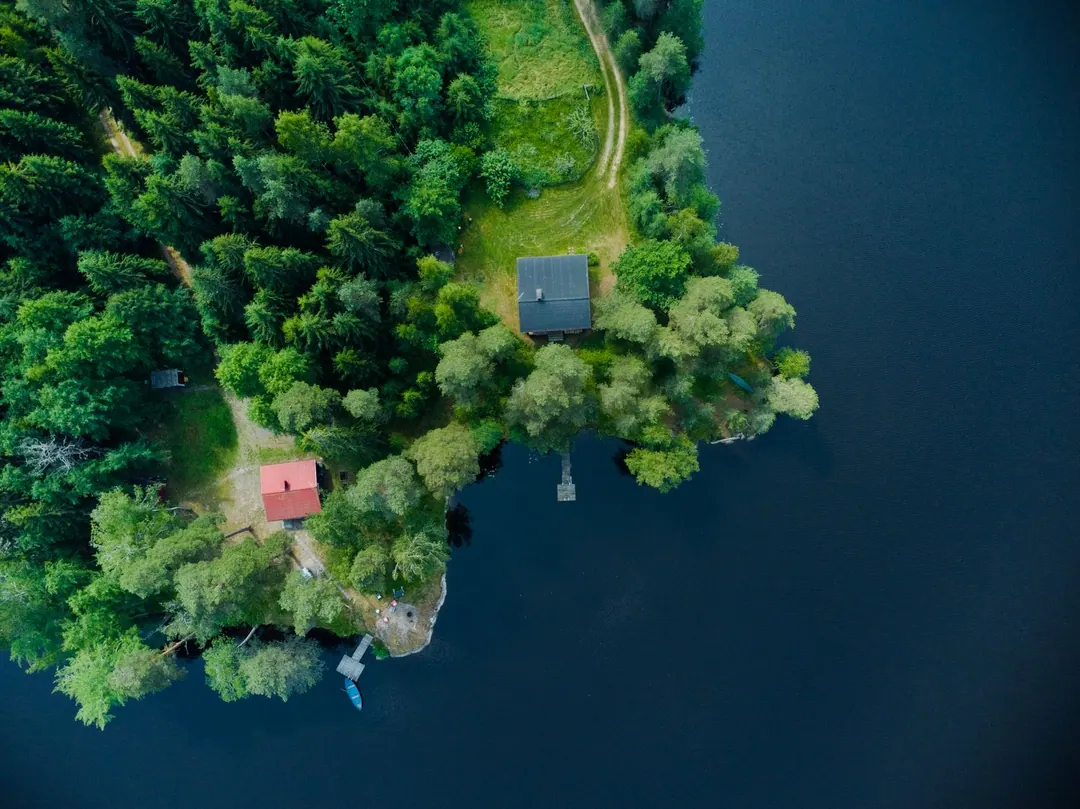 mökki - An aerial view of two small cottages on a wooded lakeshore, with jetties and a rowing boat at the water's edge
