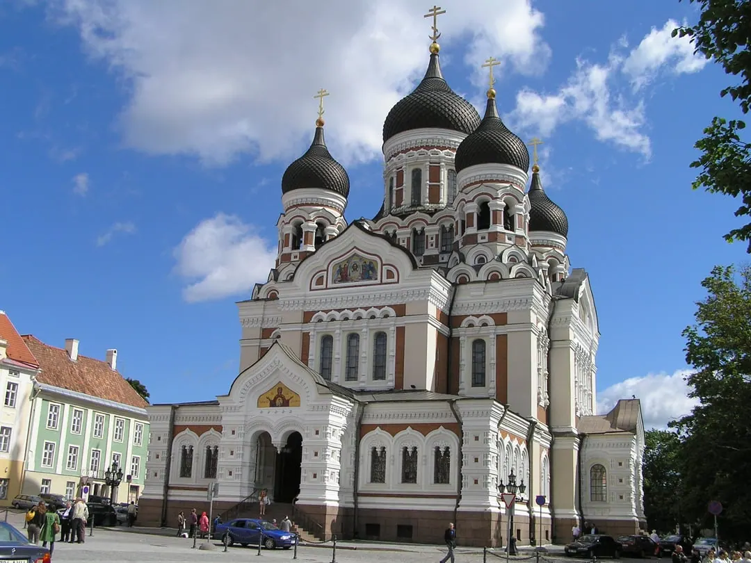 A white and pale orthodox cathedral with five black onion domes, behind a low fence and trees