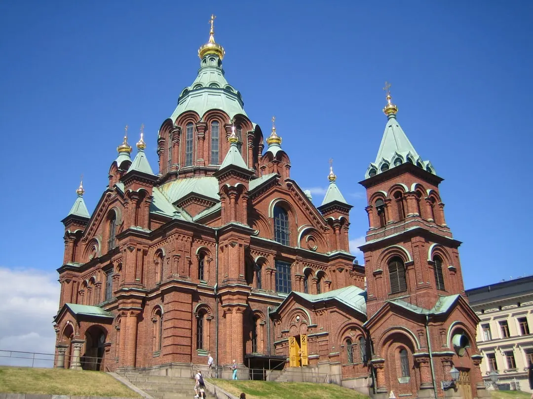 A red brick orthodox cathedral with green copper roofs and gold onion domes, on a rocky rise