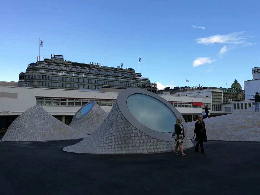A paved square with several smooth concrete mounds rising from it, each with a round skylight set into the top