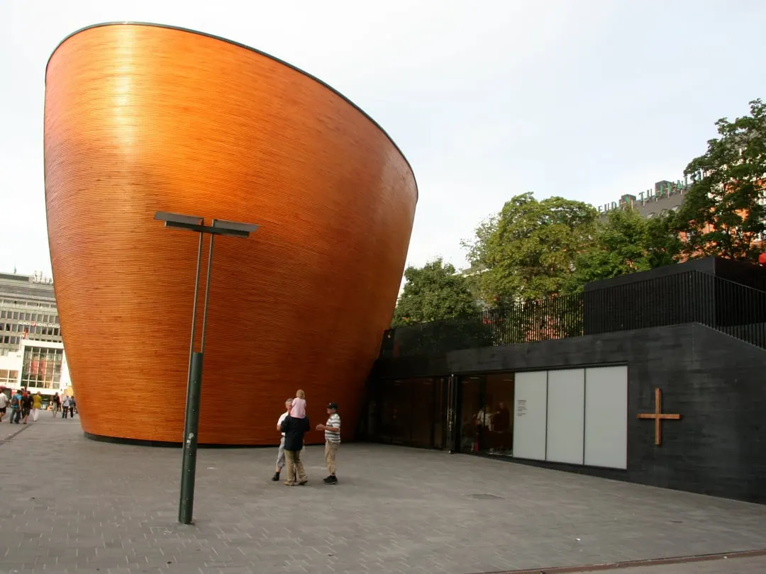 A curved windowless building clad in narrow battens of pale timber, shaped like a rounded bowl, on a city square