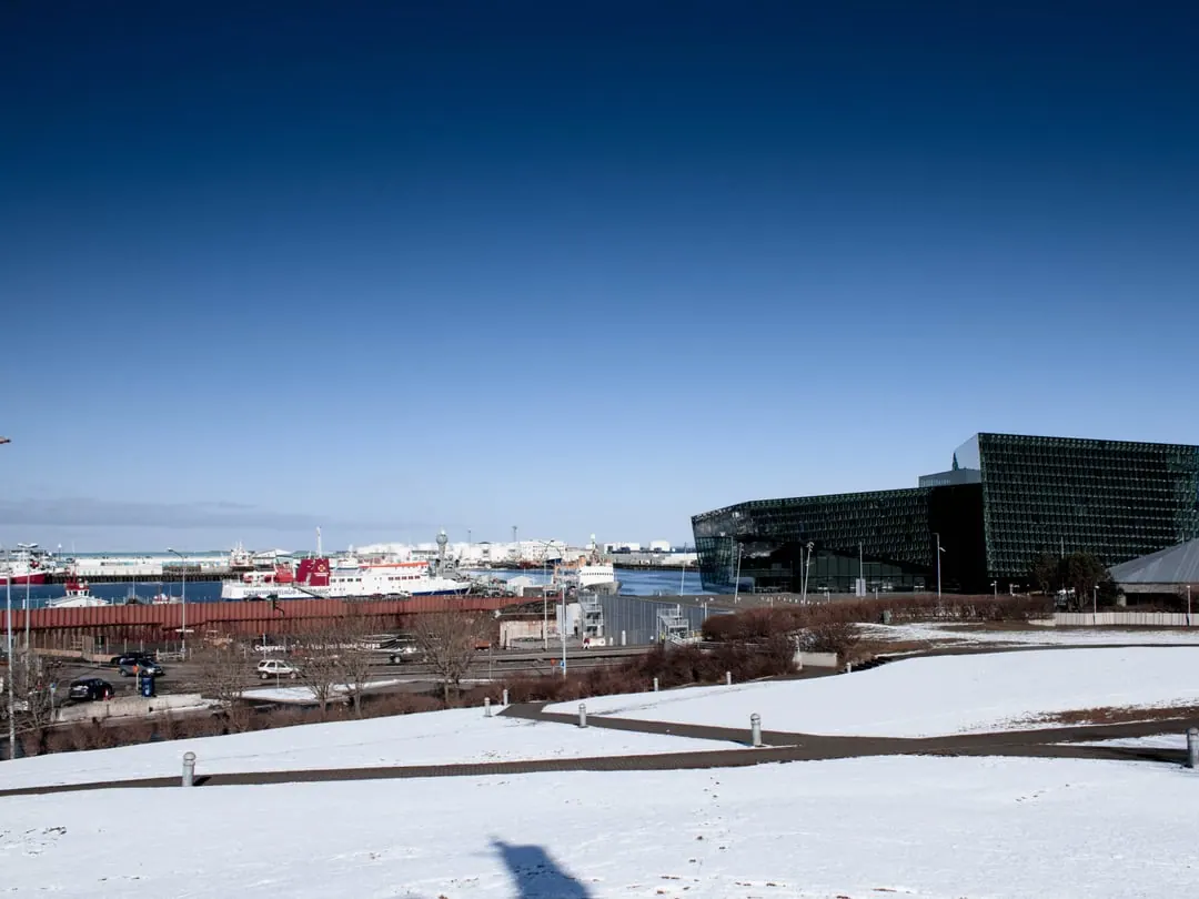 A dark glass concert hall with a faceted honeycomb facade, standing on a bare waterfront under a wide sky