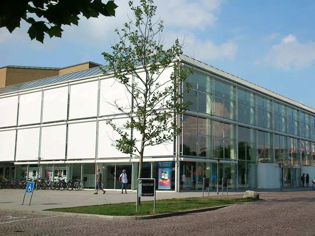 A long low concert hall in a grid of glass and pale frame, with trees in front of it