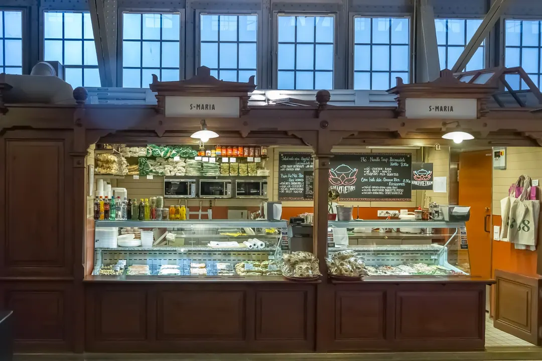 A wooden delicatessen stall inside a market hall, with a glass counter of prepared foods and a lit display above it