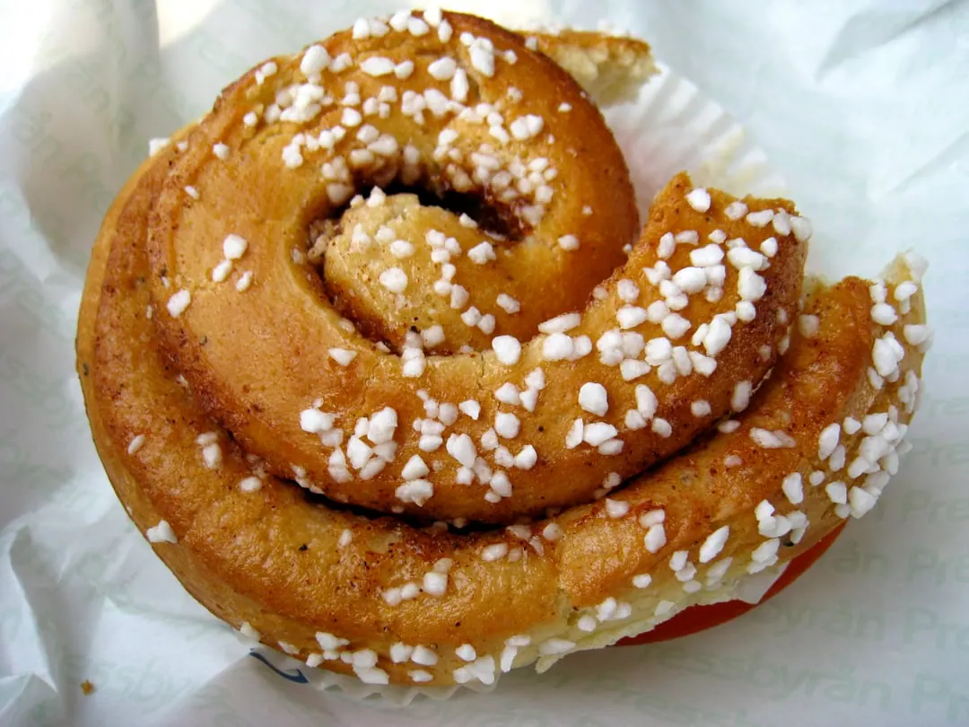 A single cinnamon bun coiled in a neat round spiral and scattered with white pearl sugar, in a paper case