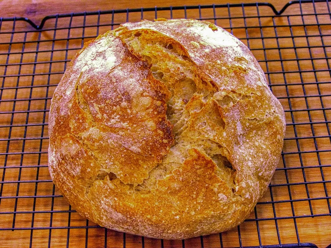 A round golden-crusted wheat sourdough loaf scored across the top, cooling on a wire rack