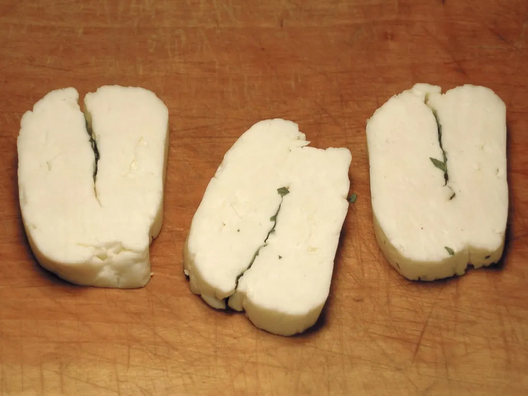 Three pale rectangular blocks of raw halloumi cheese on a wooden board, each split along one side