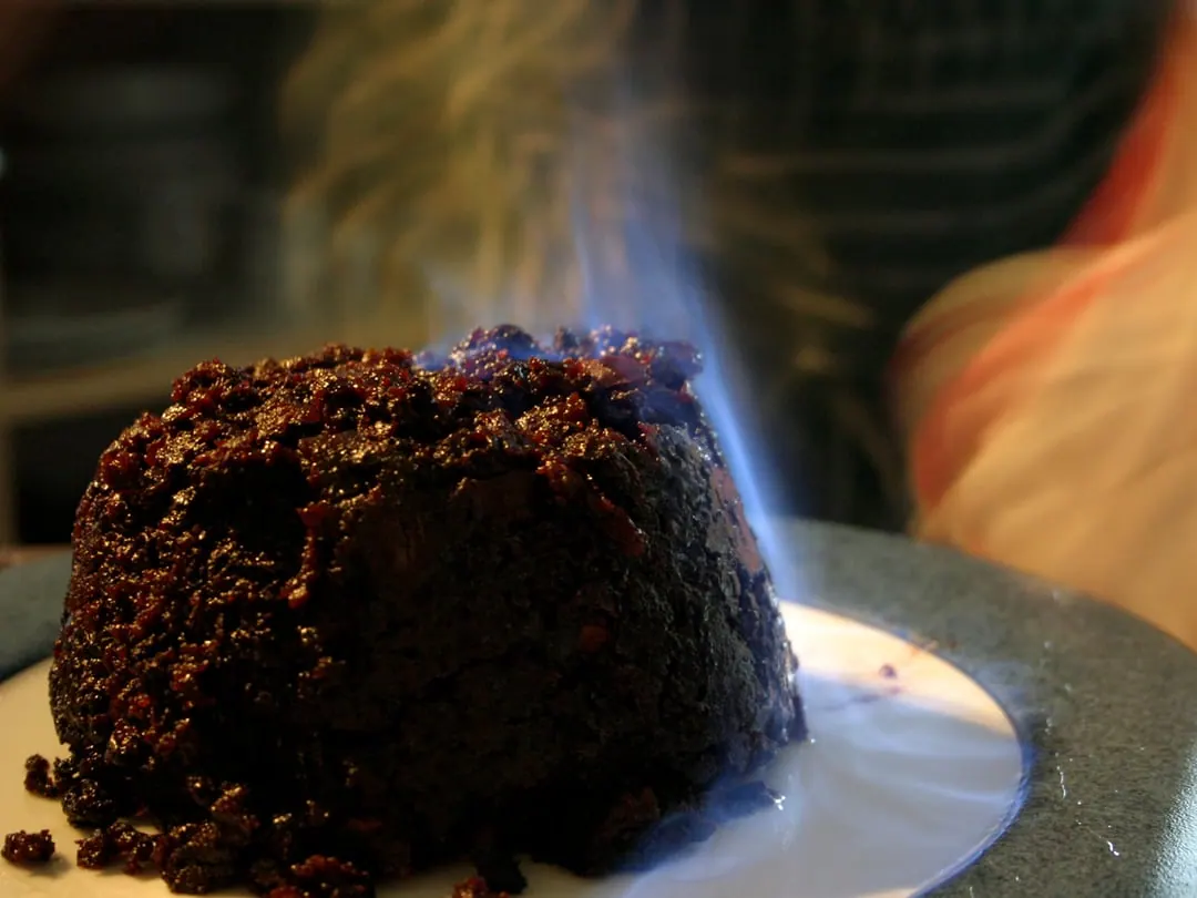 A dark domed steamed pudding alight with pale blue burning brandy, in a dish of cream