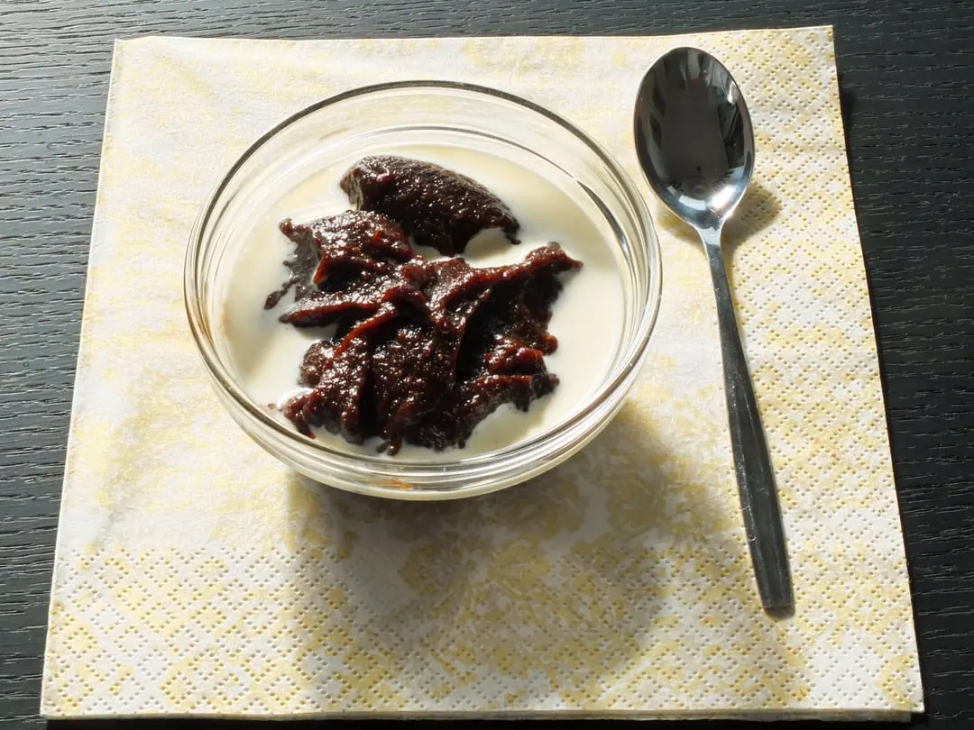 A glass bowl of very dark brown malted rye pudding with a spoon beside it on a napkin
