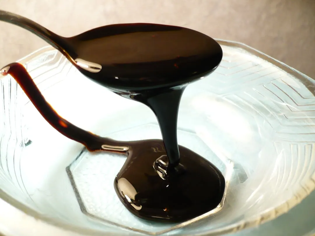 Dark molasses pouring off a spoon into a glossy pool on a white plate