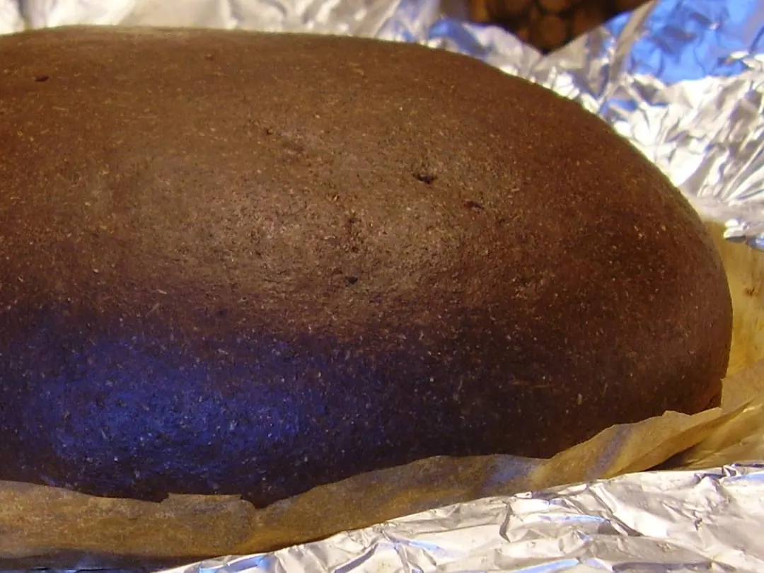 A large smooth brown rye loaf with an unscored crust, resting on foil