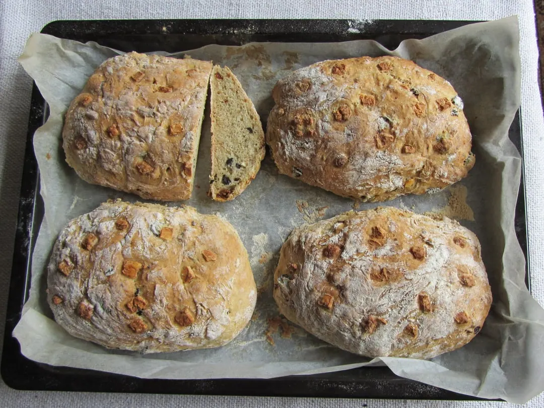 Four round seeded rye loaves on baking paper in a tray, one cut open