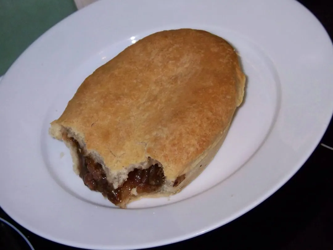 A pale short-crust pie on a white plate, broken open to show a dark meat filling