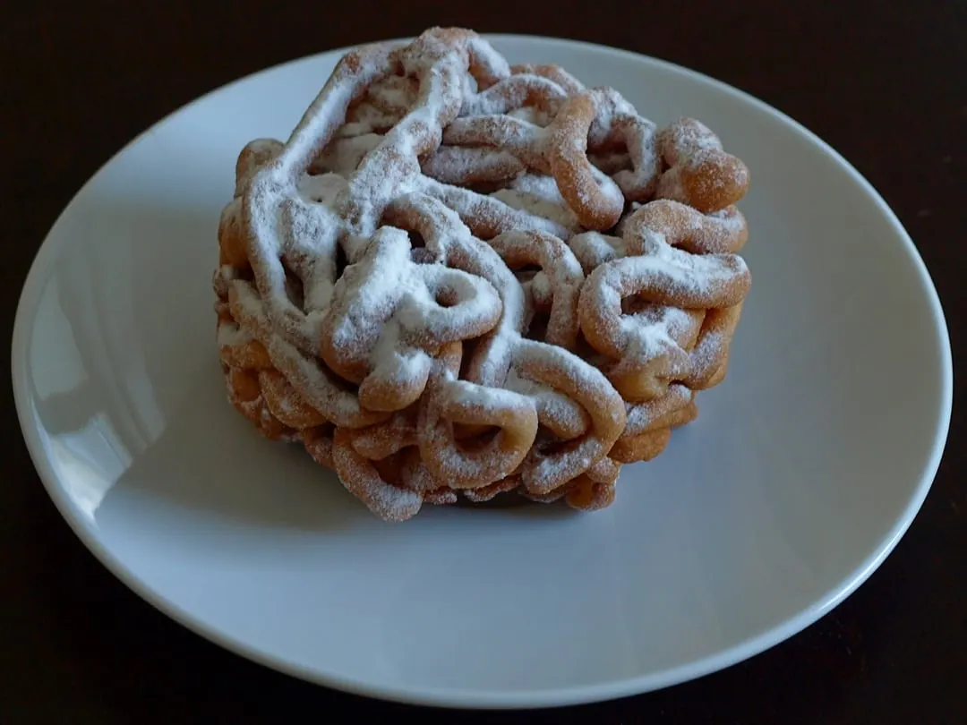 A tangled dome of thin fried batter loops buried in icing sugar, on a white plate