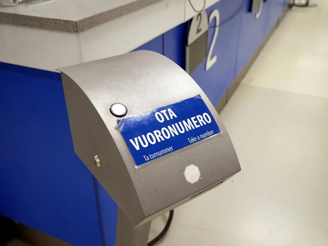 A ticket dispenser on a blue counter, its label reading OTA VUORONUMERO in Finnish, Ta turnummer in Swedish and Take a number in English