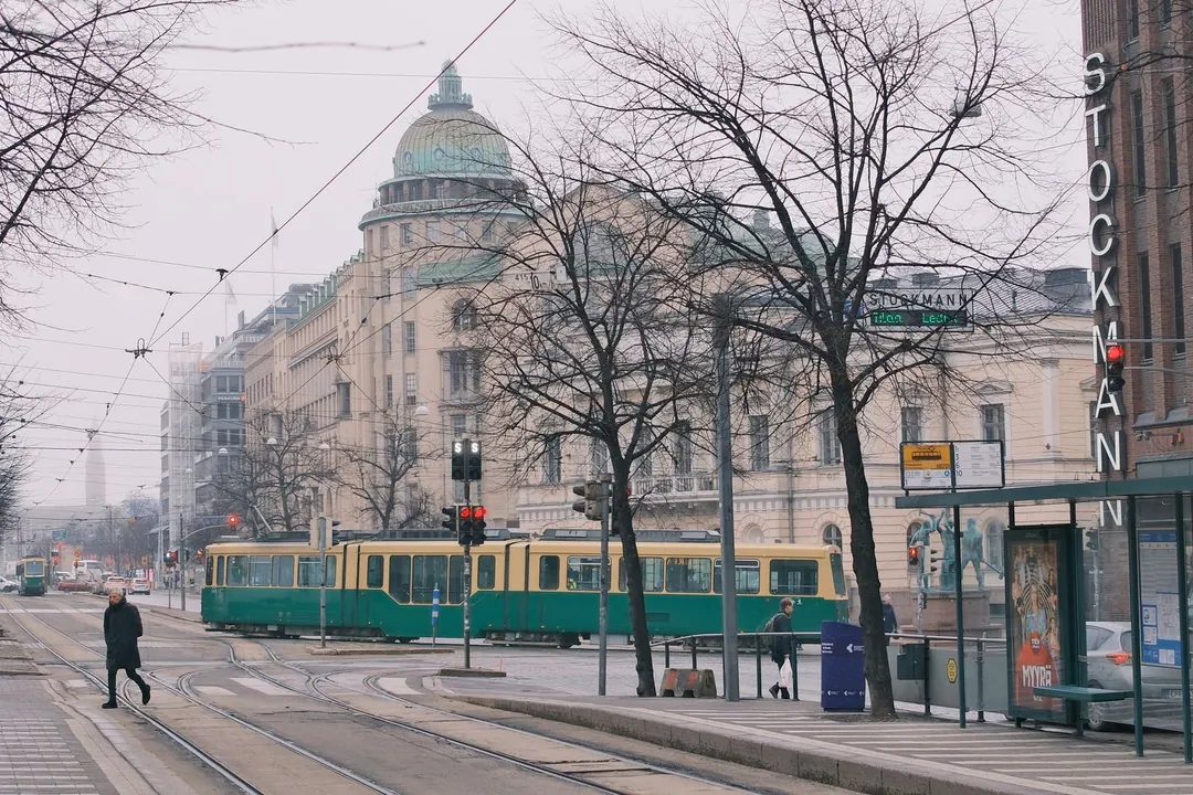 ratikka - A green and cream tram stopped at a Helsinki junction below the vertical Stockmann sign, bare trees and flat grey light