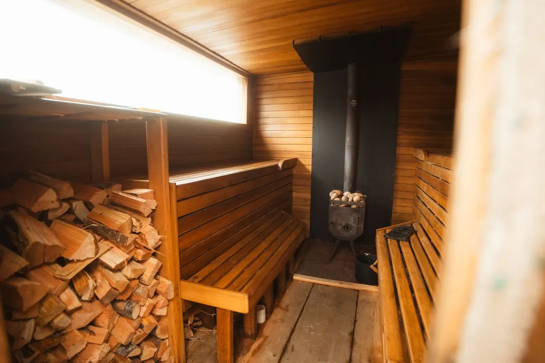 sauna - A wood-fired sauna with a stacked woodpile, two tiers of wooden benches and a black stove with stones piled on top